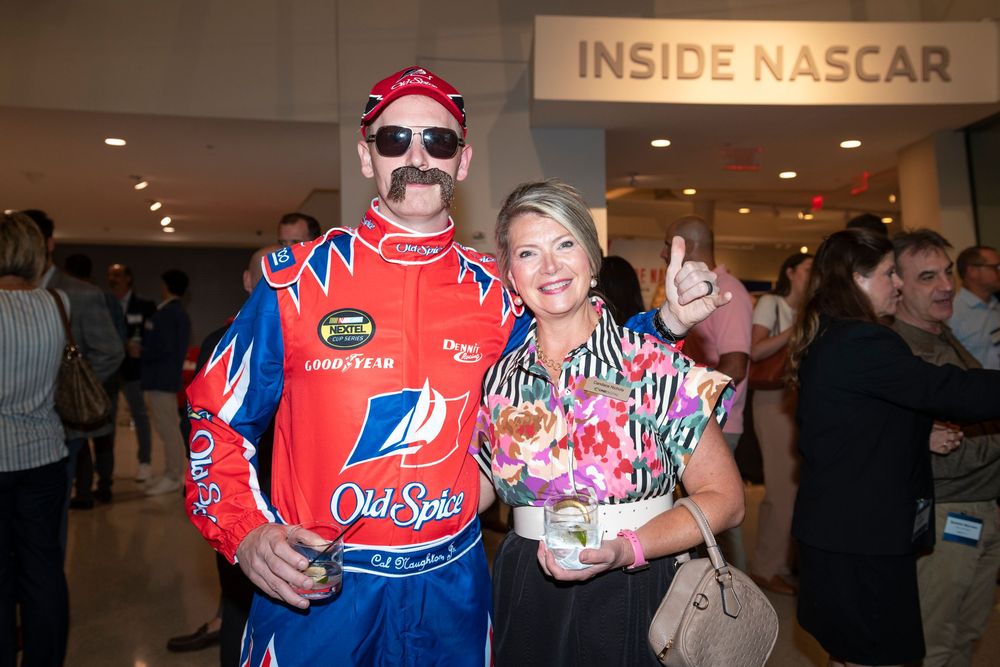A NASCAR driver in a red racing suit poses with fans at a meet and greet event.