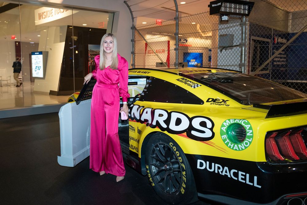 A bright pink mascot character interacts with a yellow race car in what appears to be an indoor venue or showroom.