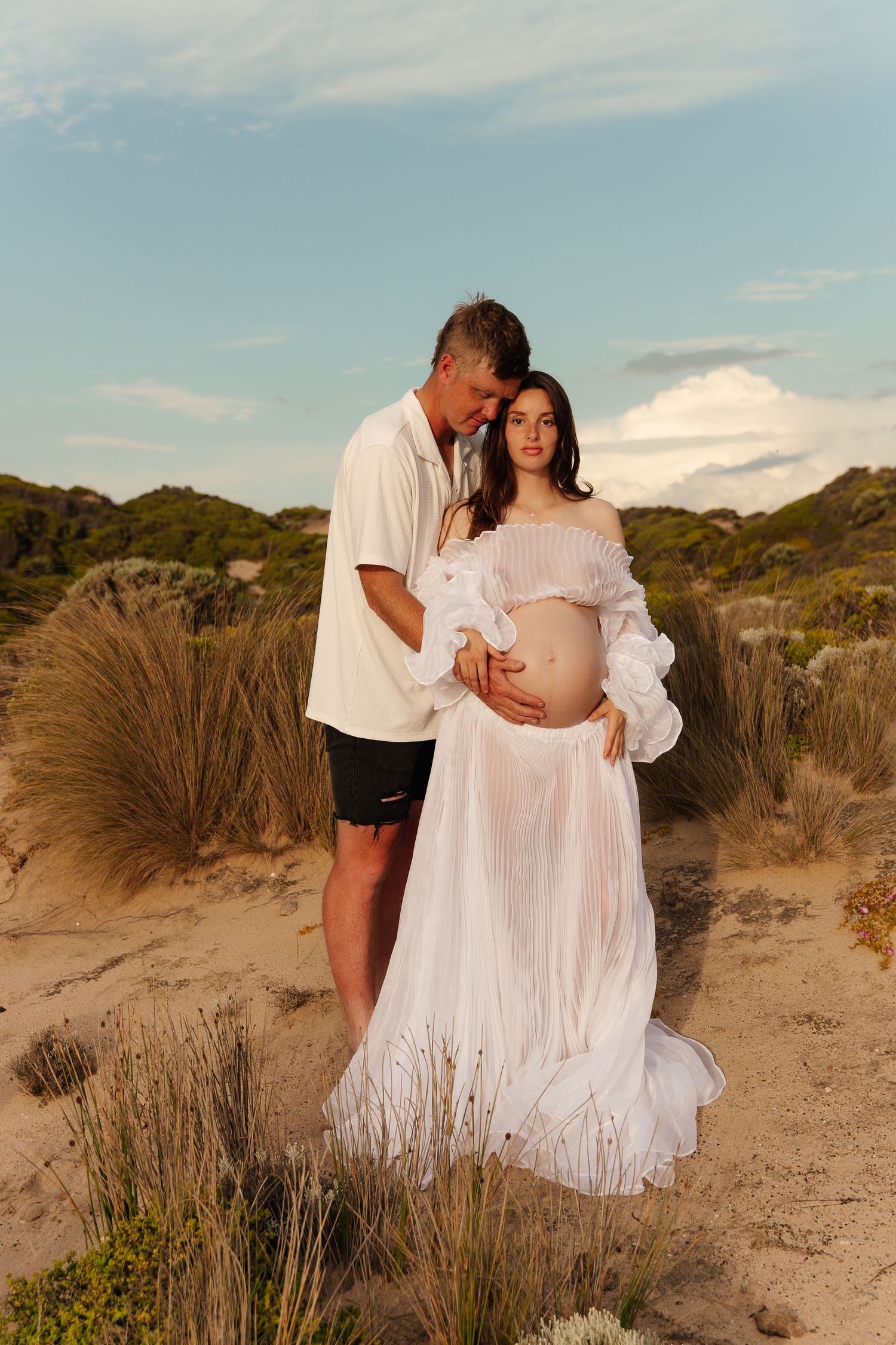 Couple in white attire embracing on a sandy beach with coastal vegetation and dunes at sunset.