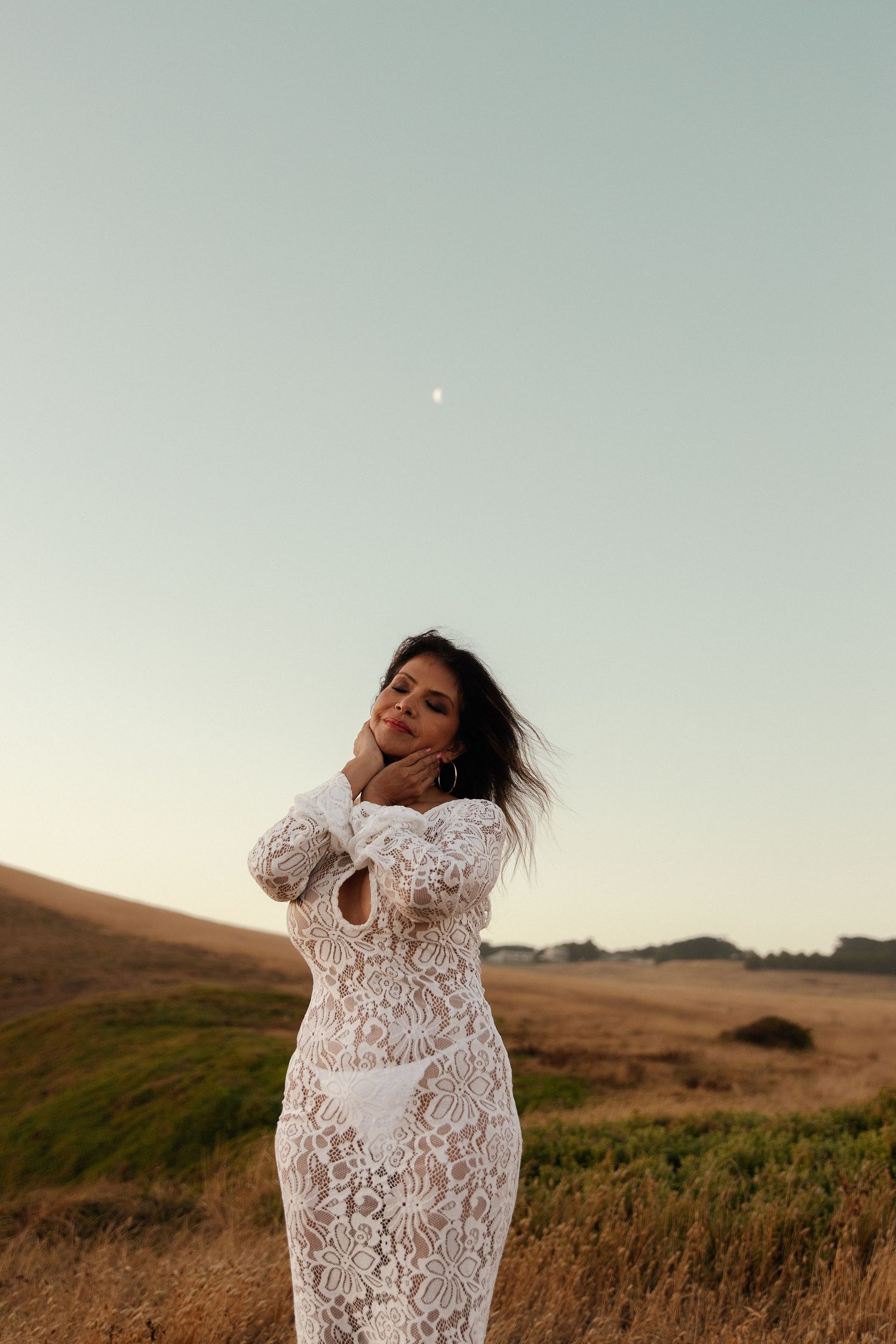 Person in white lace dress poses against sunset in grassy field with wind-swept hair.