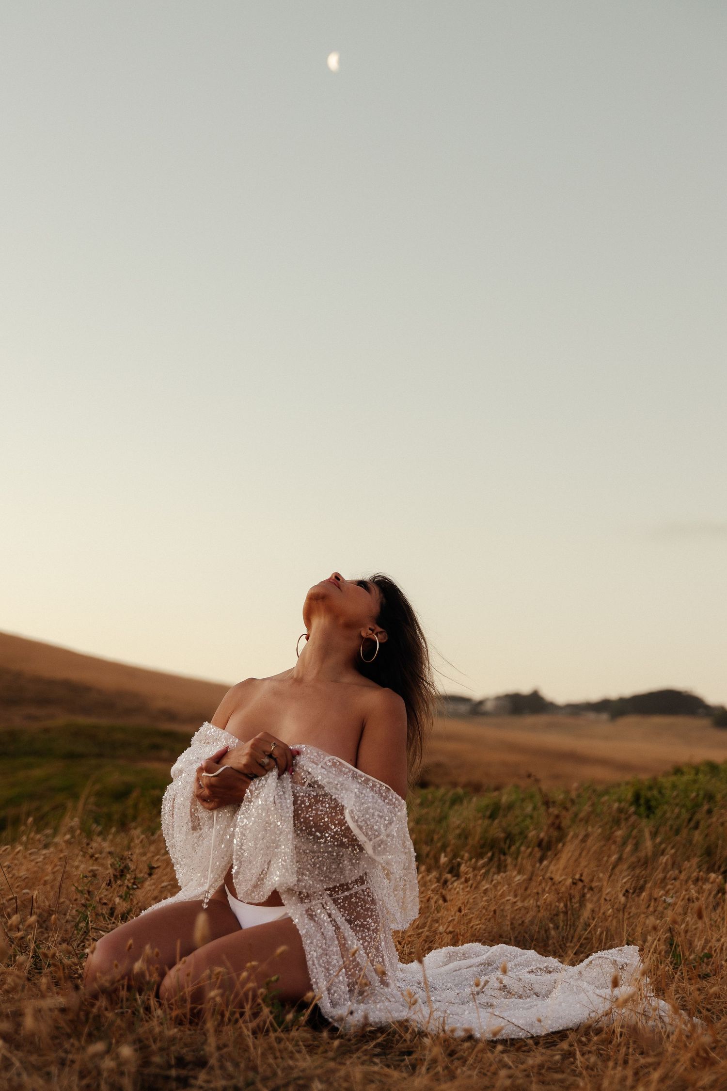 A person in a white lace dress sits in a golden field at sunset with the moon visible in the pale sky.