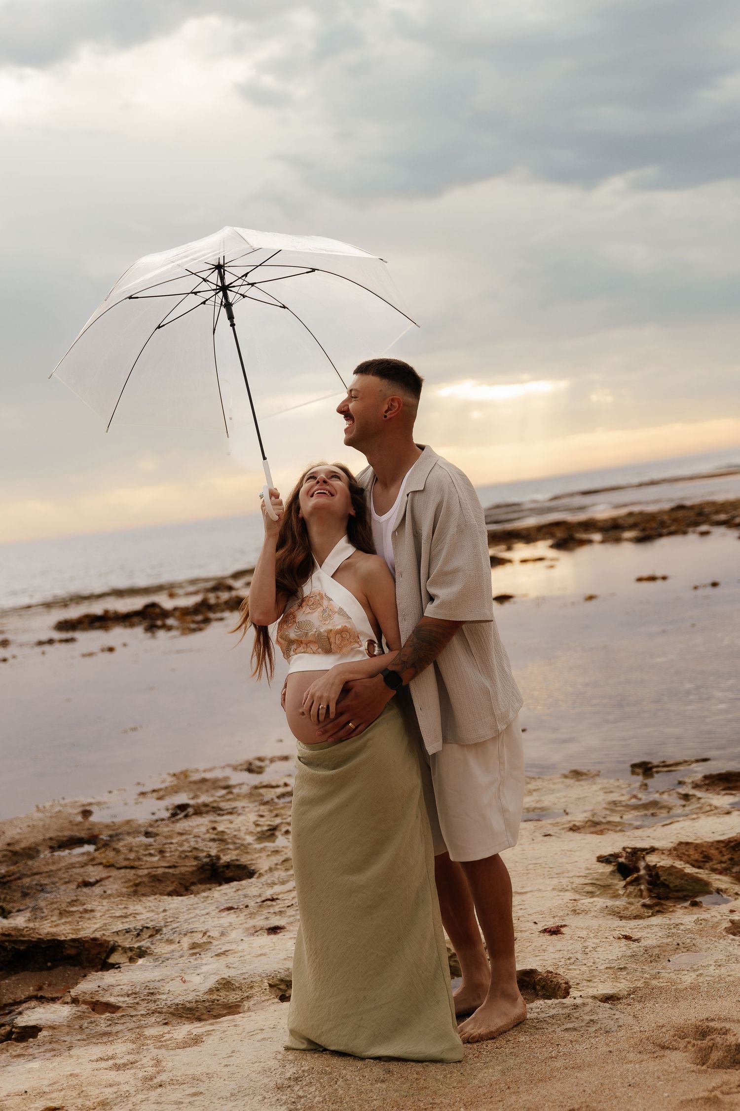 A couple shares intimate moments on a rocky beach at sunset under a white umbrella.