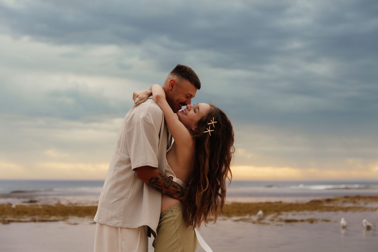 Couple embraces on sandy beach during cloudy sunset with dramatic sky overhead.