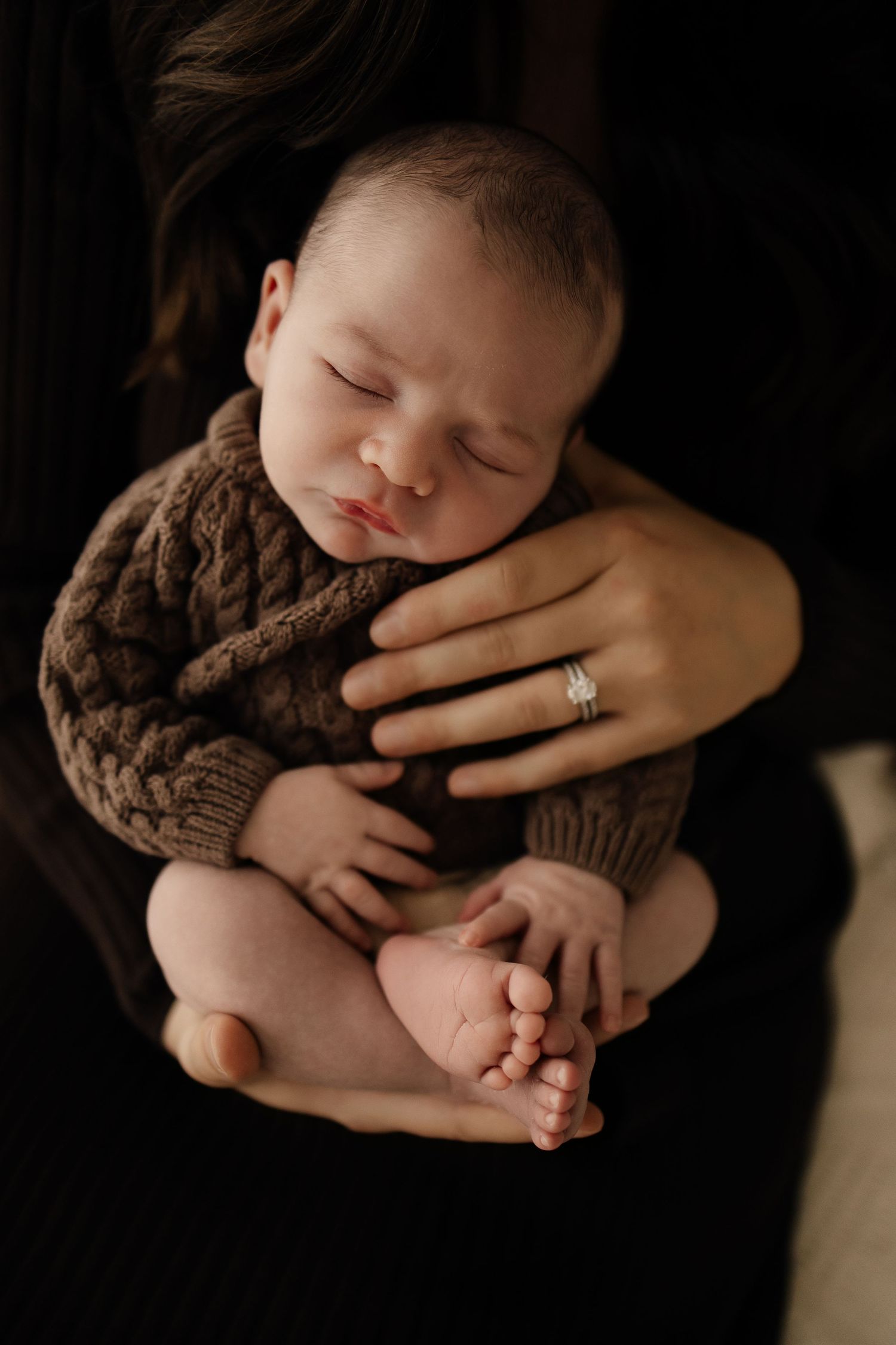 A sleeping newborn baby wrapped in a brown knit blanket is held gently while tiny feet peek out in dramatic lighting.