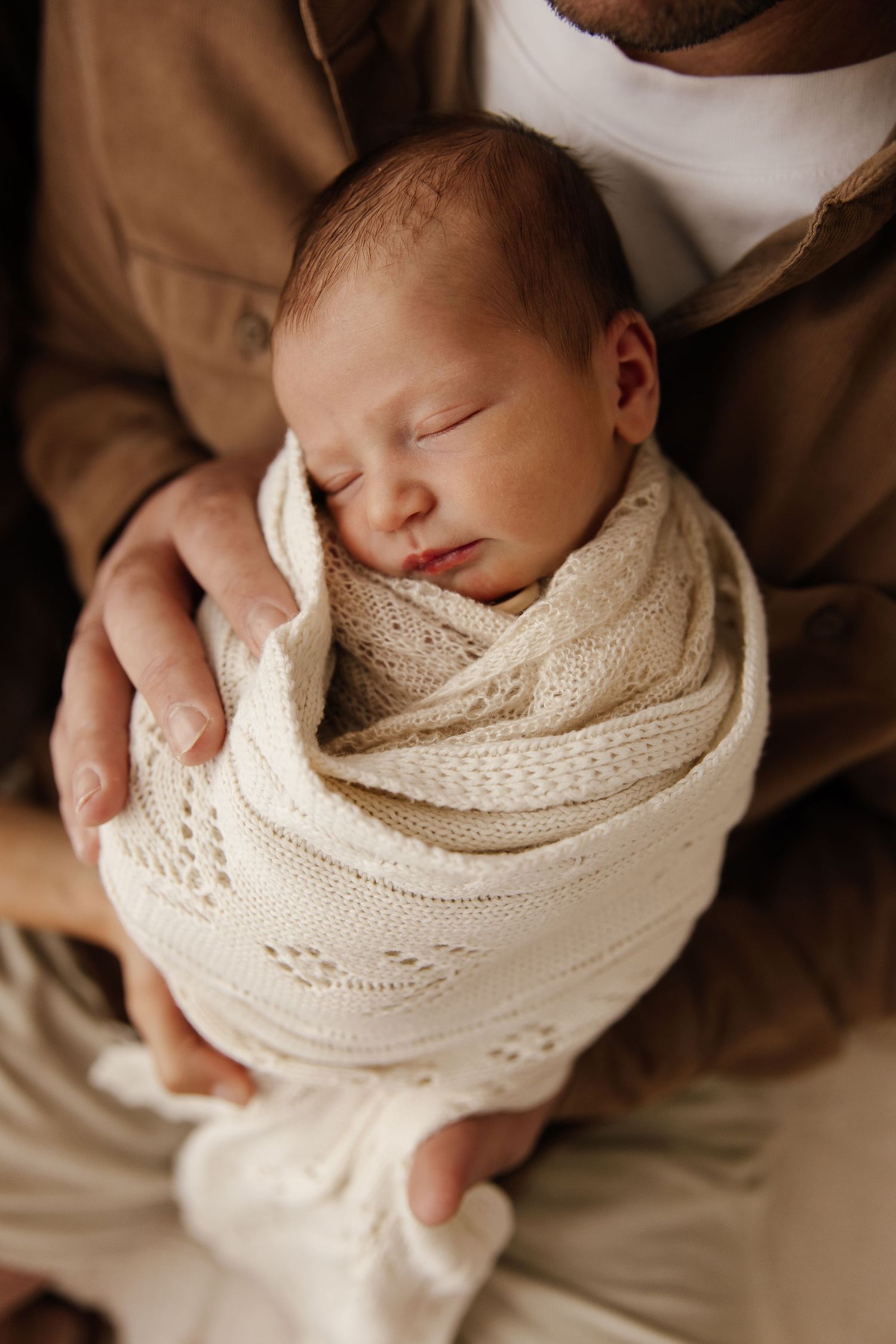 A newborn baby wrapped in a cream-colored knit blanket is held gently in caring hands.