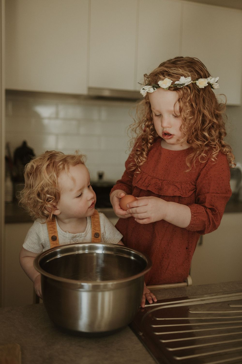 Two curly-haired children baking together in a kitchen, girl with flower crown holding an egg over a mixing bowl.