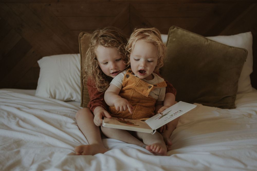 Two young siblings sit together on a white bed reading a book, with the older child lovingly holding the toddler.