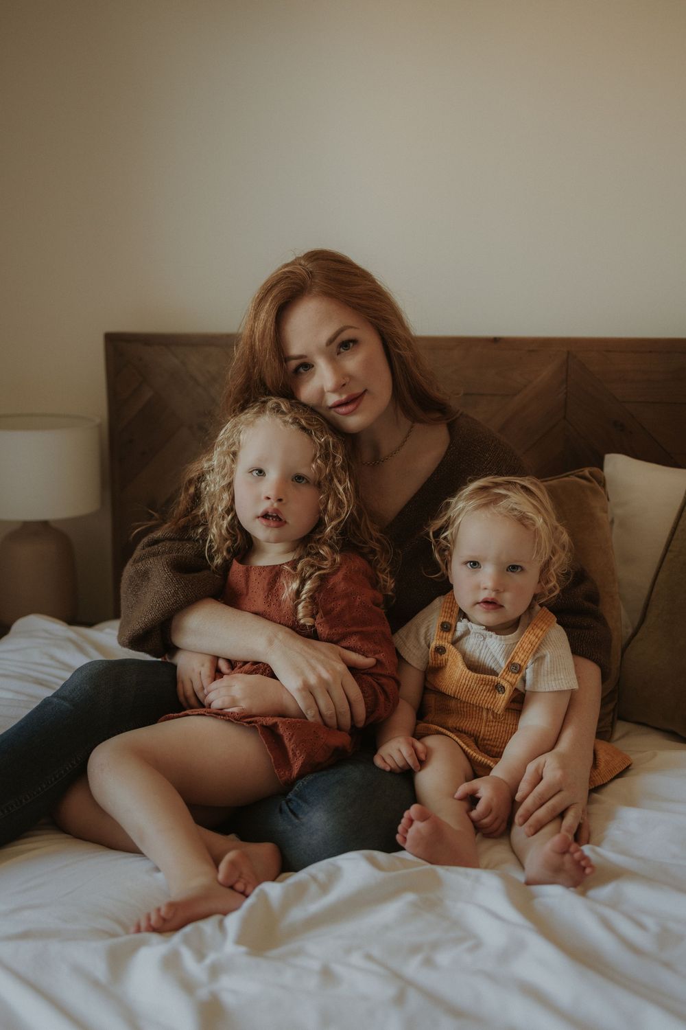 A red-haired mother sits on a white bed with her two young curly-haired children in warm-toned outfits.