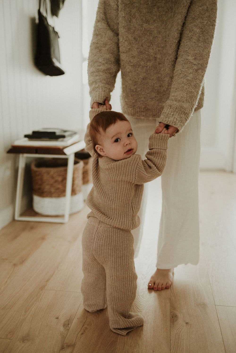 A baby in a beige knit outfit stands on hardwood floors while someone in matching attire stands nearby.