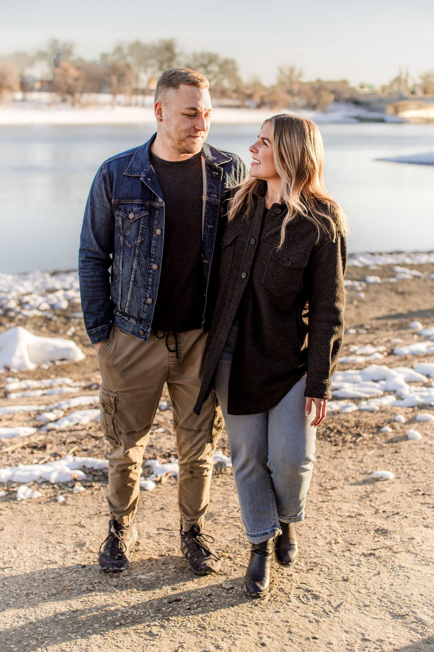 A couple walks hand-in-hand near a snowy lake, smiling at each other in winter sunshine.