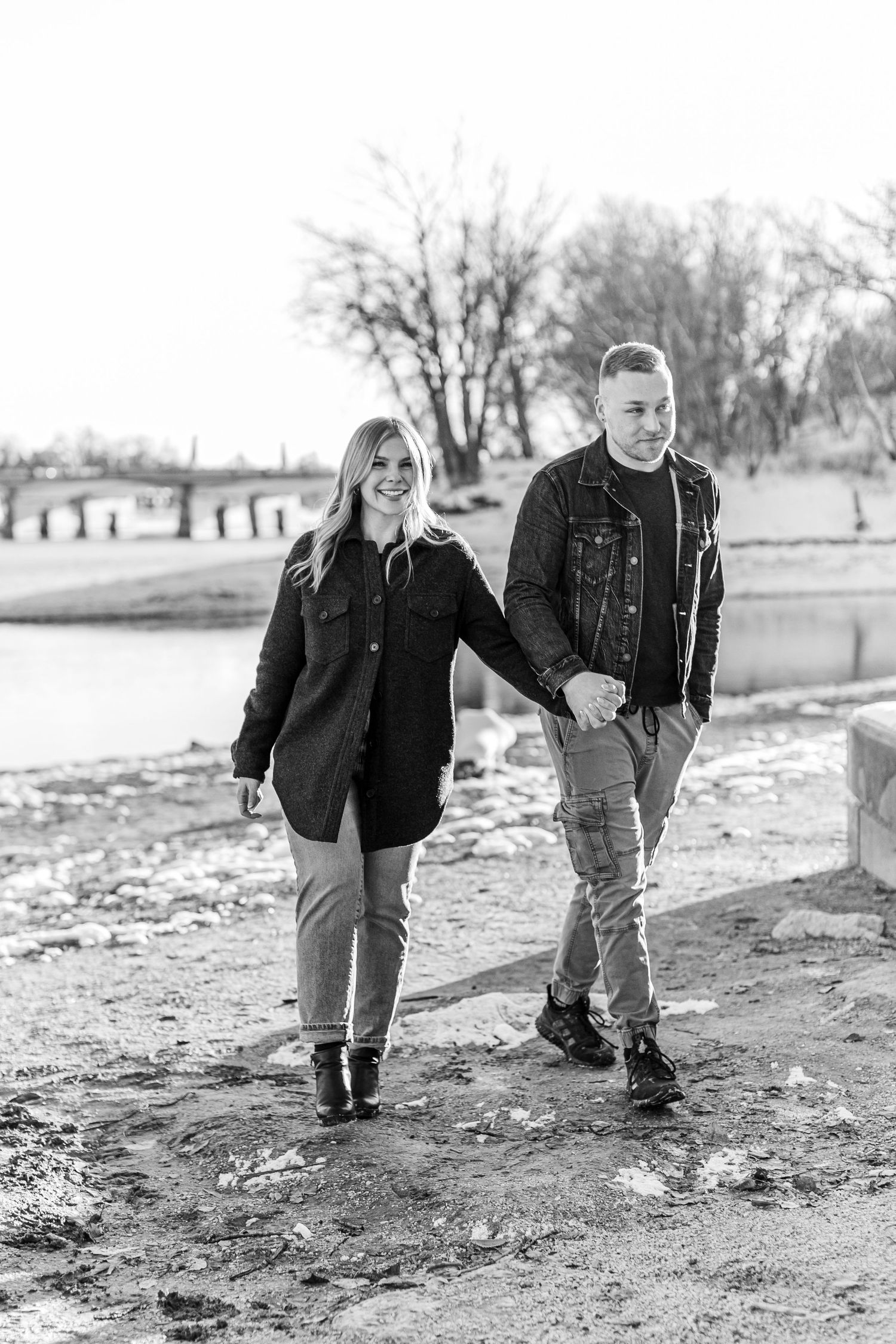 A black and white photo of a couple holding hands and walking near a winter lake.