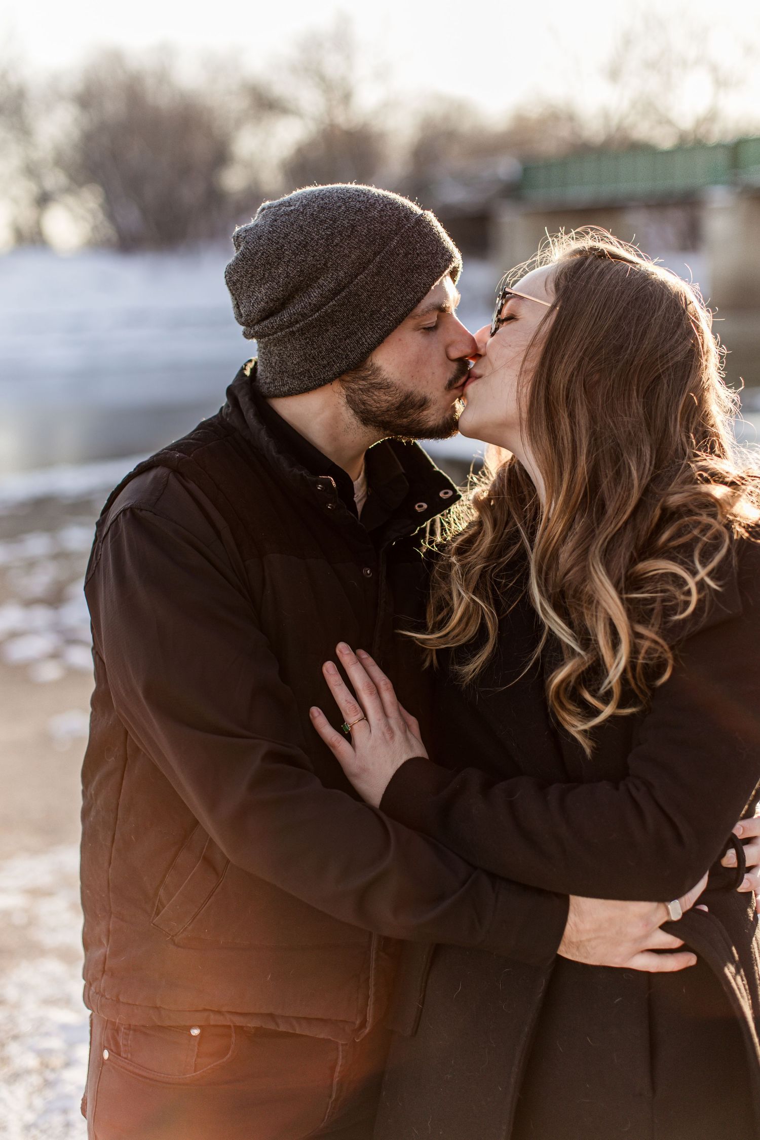 A couple shares a romantic kiss near a frozen river in warm winter sunlight.