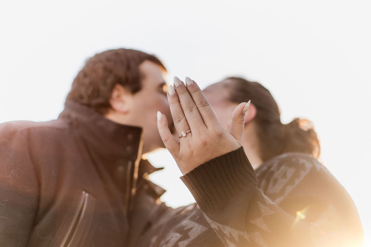 An engaged couple kisses while the woman displays her diamond engagement ring in sunlight.
