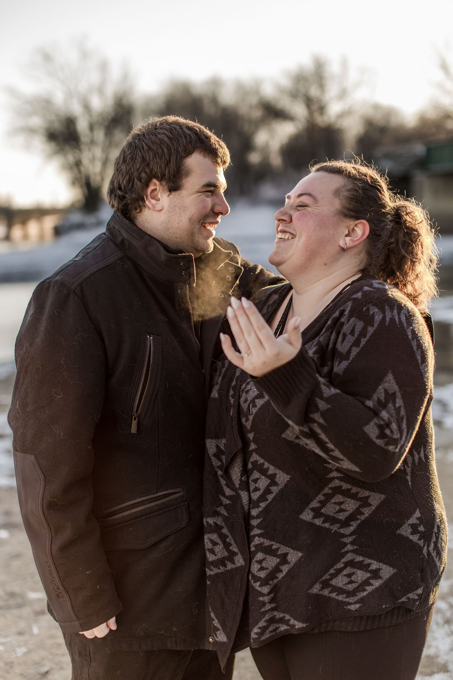 An engaged couple laughs together outdoors in winter light, the woman displaying her ring.