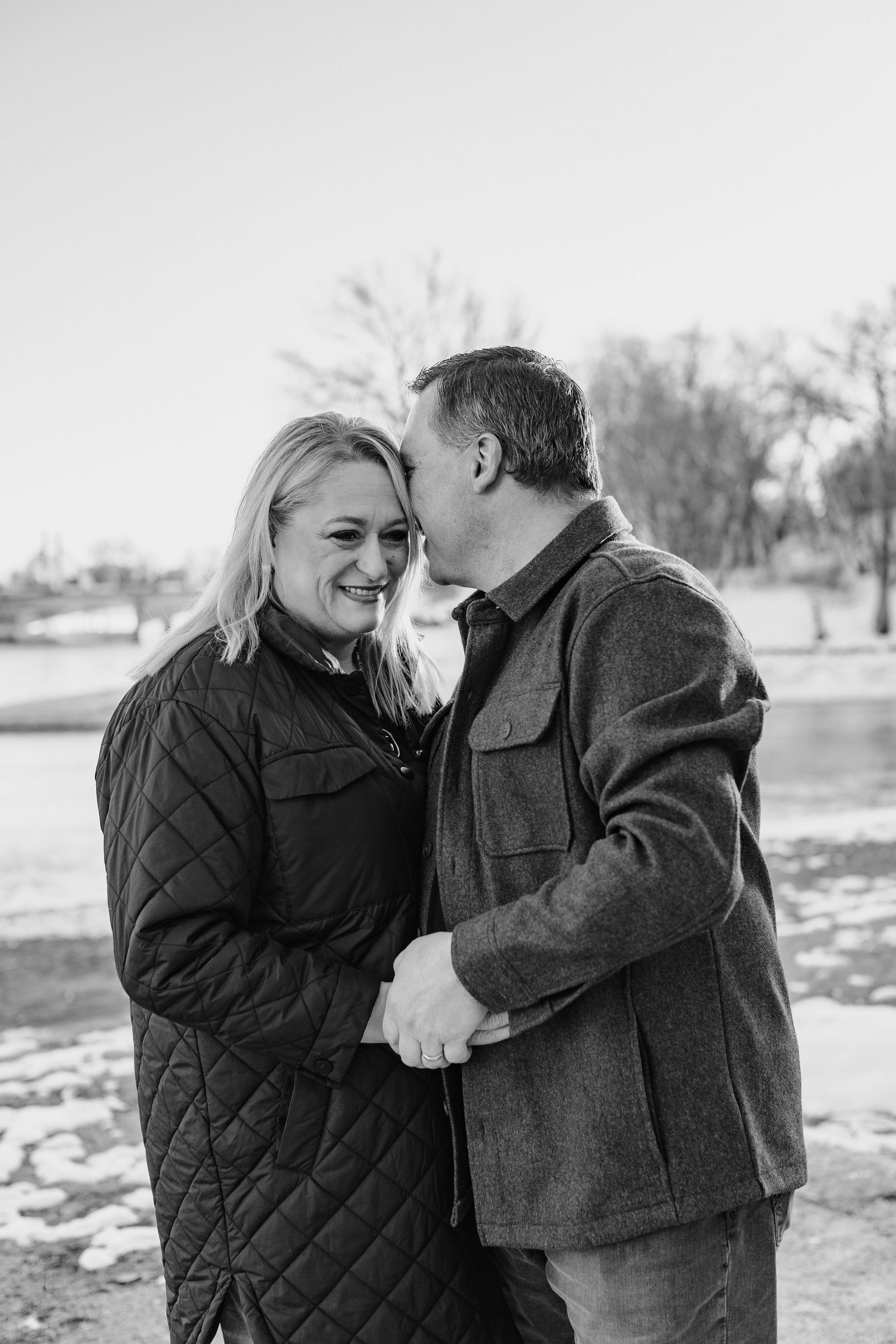 A black and white photo of a man kissing a smiling woman on the cheek near a lake.