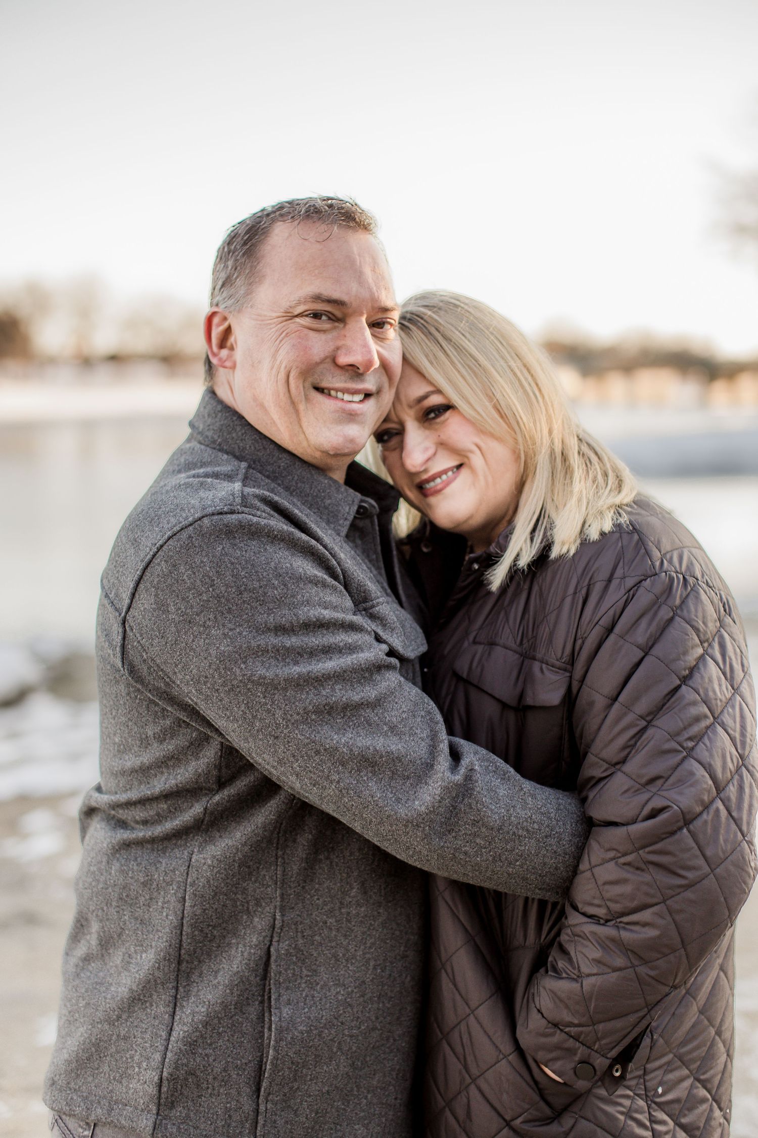 A smiling couple embraces near a frozen lake, wearing winter coats in golden hour light.