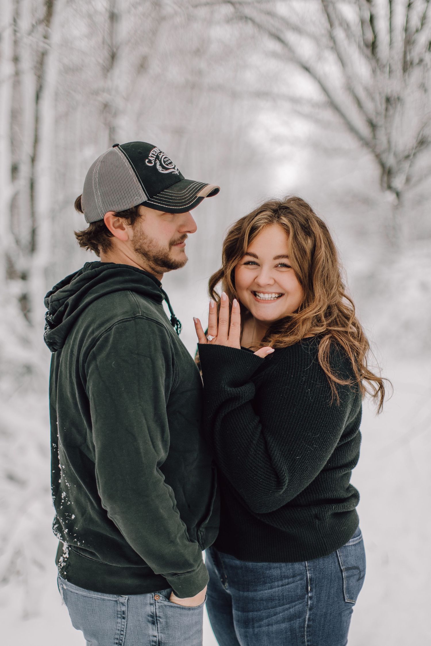 A couple embraces in a snowy winter forest, the woman smiling and showing her engagement ring.