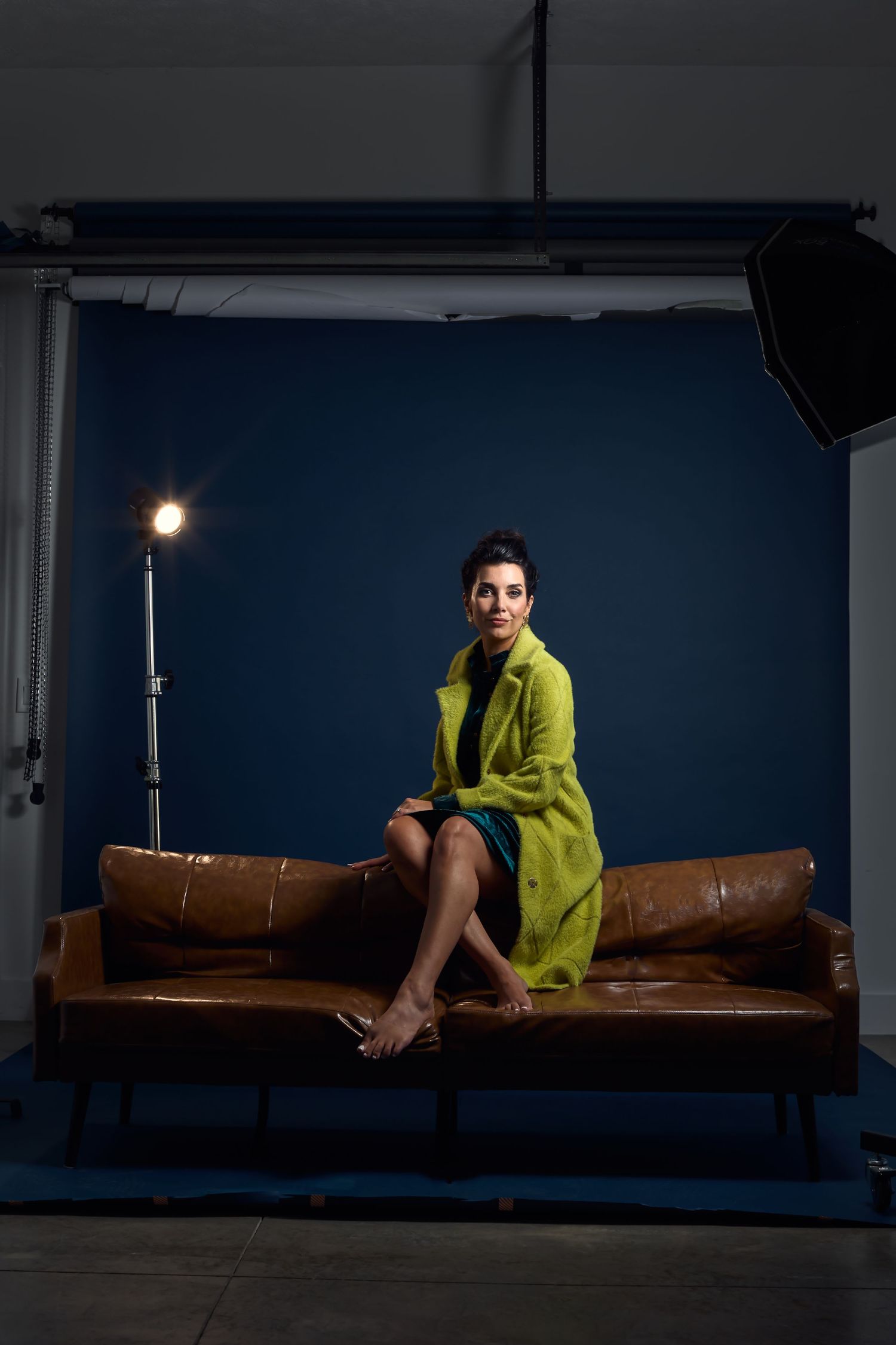 Stylish portrait in bright yellow dress seated on brown leather couch in studio setting.