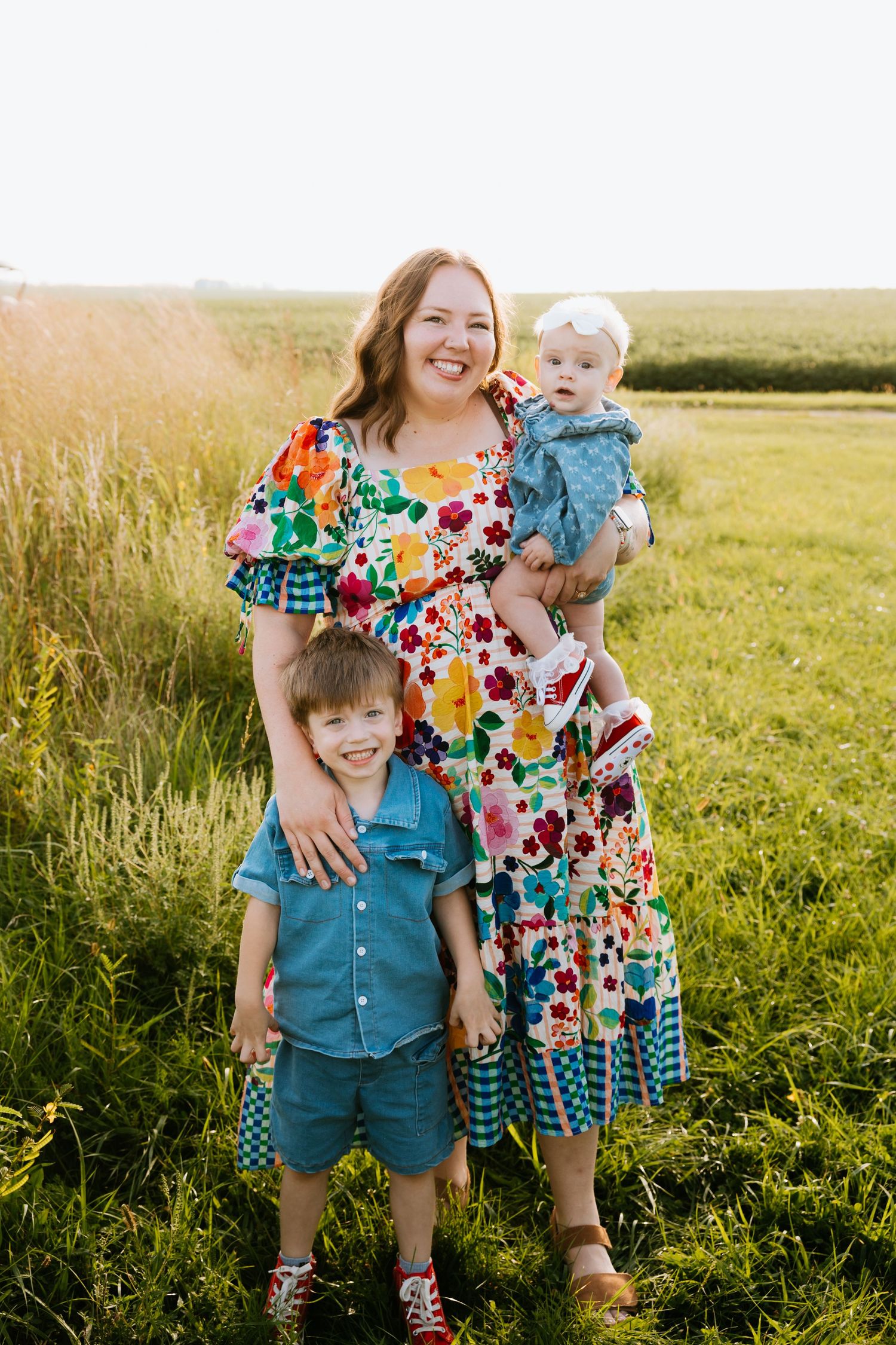 Radiant mother in colorful floral dress holds baby and hugs young son during outdoor family portrait.