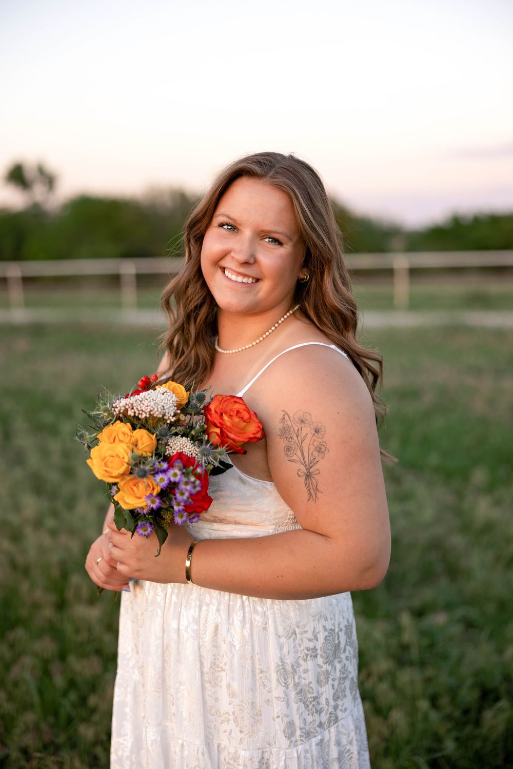 Smiling bride in white lace dress holding colorful bouquet of roses outdoors at sunset with green field background.