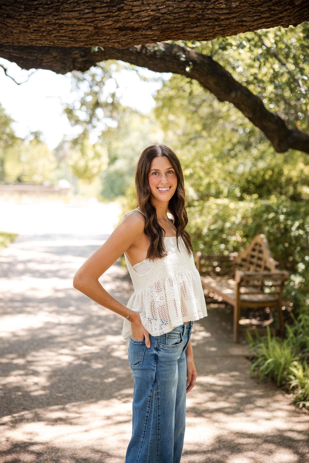 Smiling brunette woman in white lace top and wide-leg jeans posing outdoors under oak tree on sunny day.