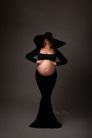 Zwangere vrouw in zwarte jurk en grote hoed  tijdens fotoshoot in studio