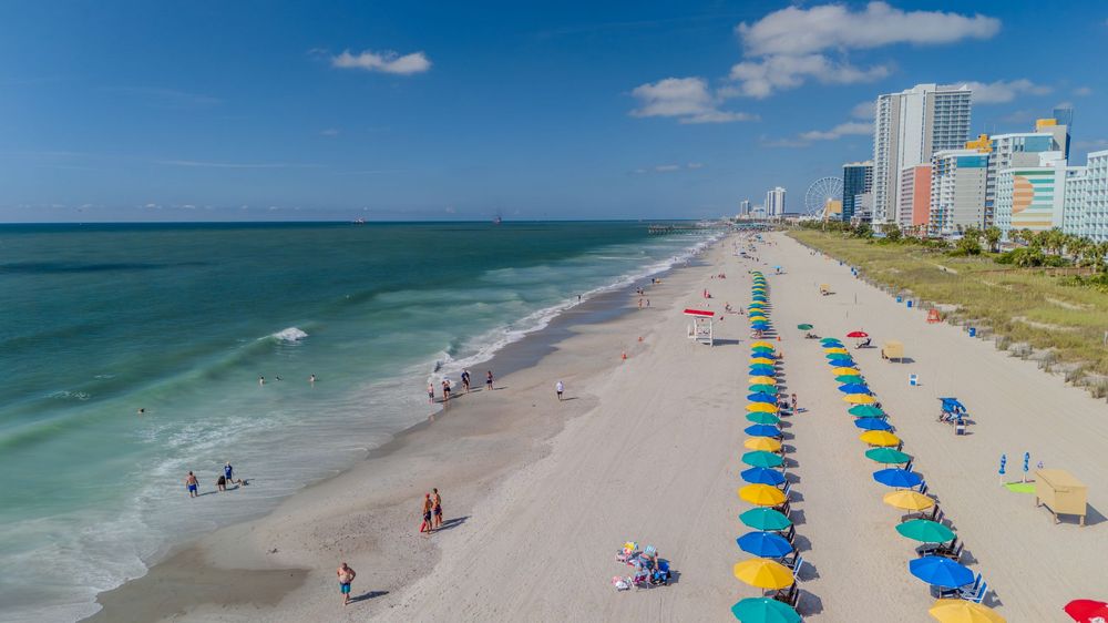 Aerial view of a sunny beach with colorful umbrellas, calm turquoise waves, white sand, and city skyline in the background.