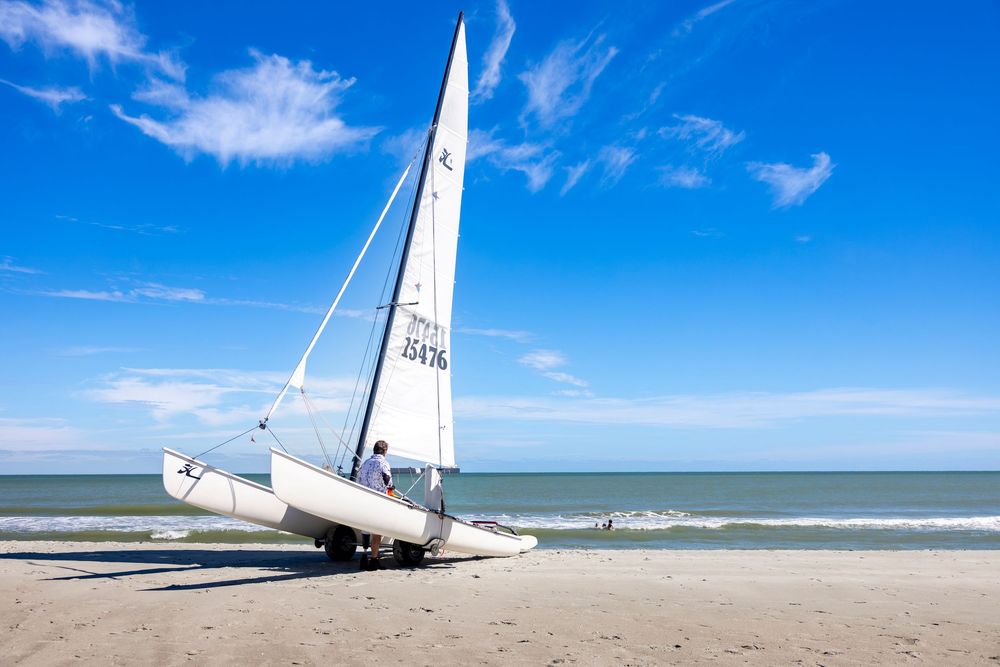 A white catamaran sailboat on a sandy beach with blue skies and calm ocean waves in the background.