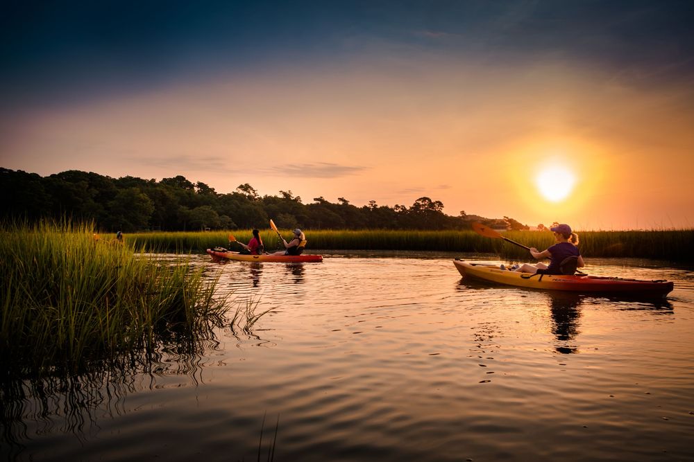 Two kayakers paddling on a calm marsh waterway at golden sunset with lush green reeds and glowing sky.