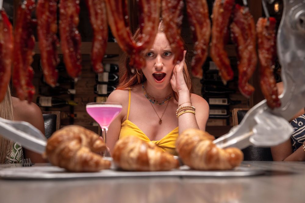 Surprised redhead woman in yellow dress with pink martini, croissants, and hanging meat in background.