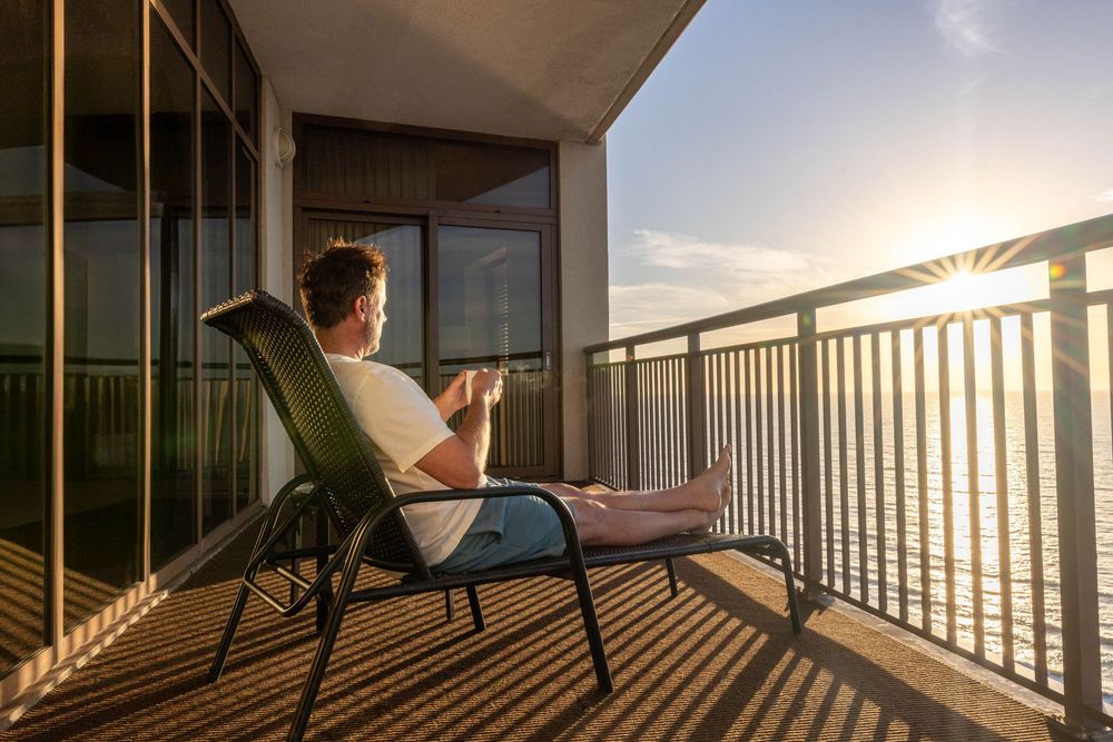 Man relaxing on a balcony chair with a coffee cup, enjoying a stunning golden sunset over the ocean.