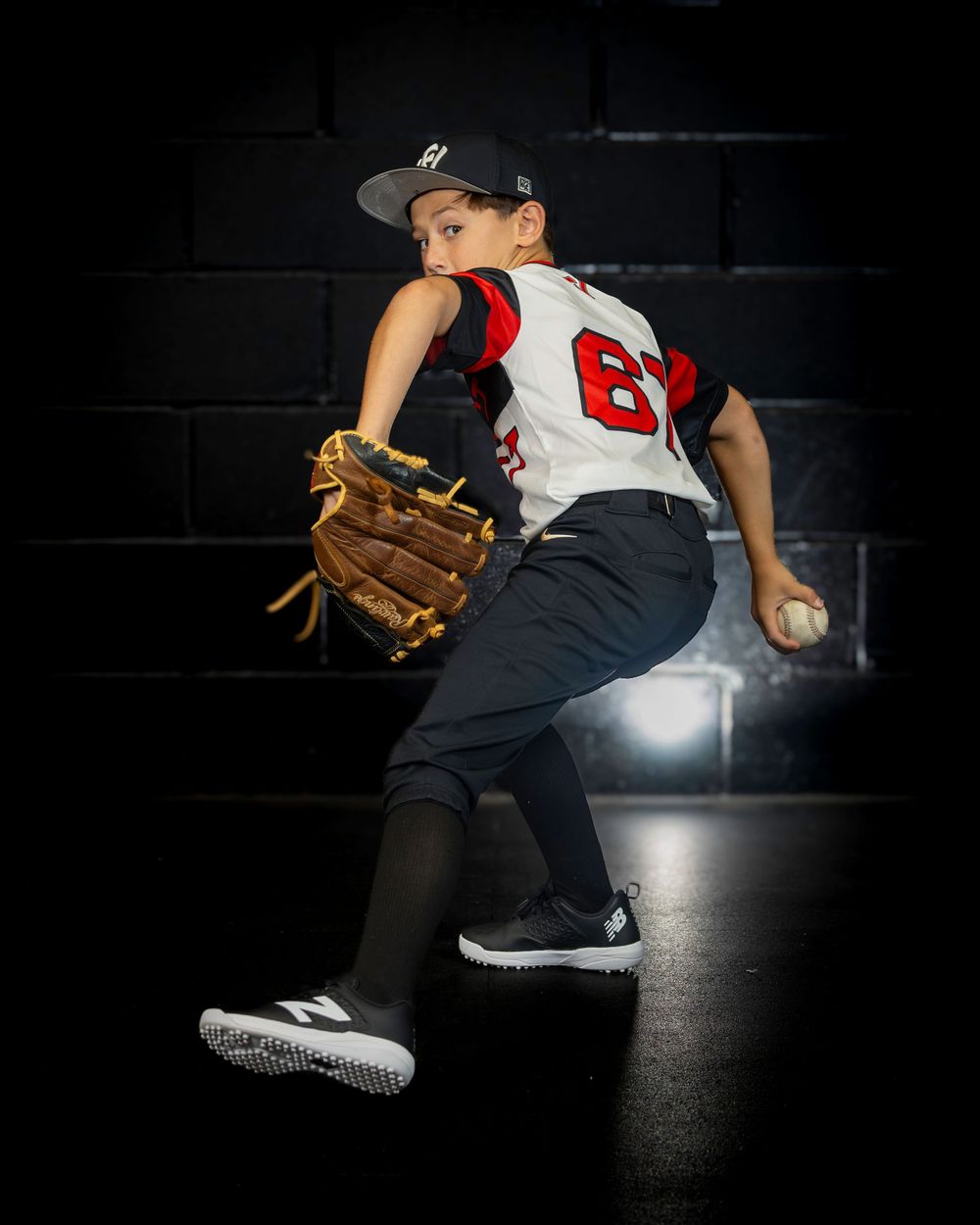 Young baseball pitcher in red and white uniform #67 winds up to throw during a dramatic dark studio photo session.