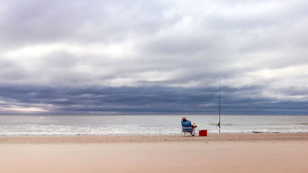 A lone fishing rod and blue chair sit on an empty beach under dramatic cloudy skies at dusk.