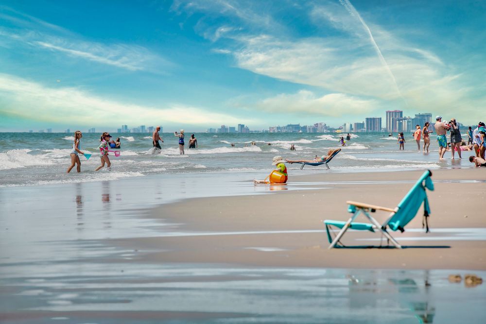 Busy beach scene with people playing in ocean waves, a teal lounge chair in foreground, and city skyline in background.