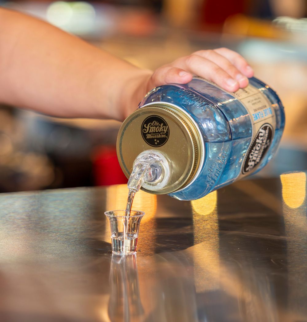 Ole Smoky moonshine being poured from a blue mason jar dispenser into a shot glass on a bar counter.