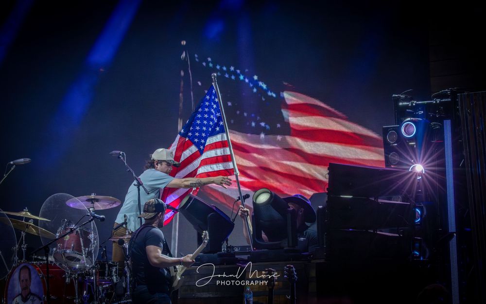 Performer waves large American flag on stage at a patriotic live concert with dramatic blue lighting and band equipment.