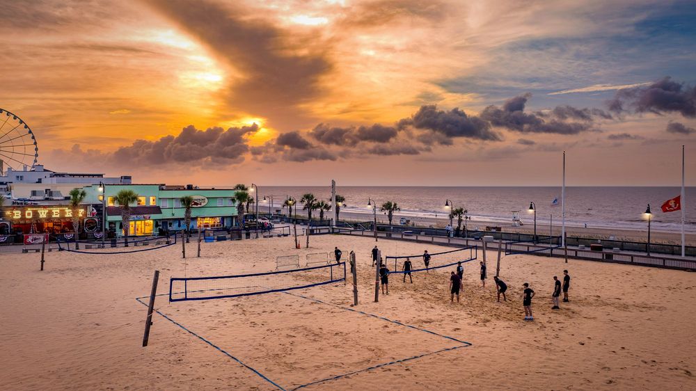 Beach volleyball players compete on sandy courts at sunset near a pier with a Ferris wheel and colorful boardwalk buildings.
