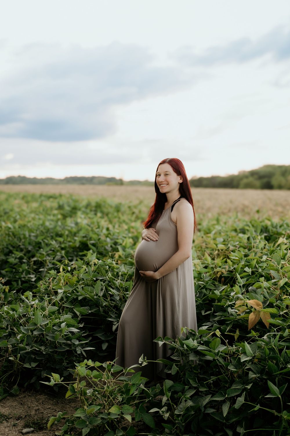Expectant mother in grey dress poses in soybean field during golden hour.