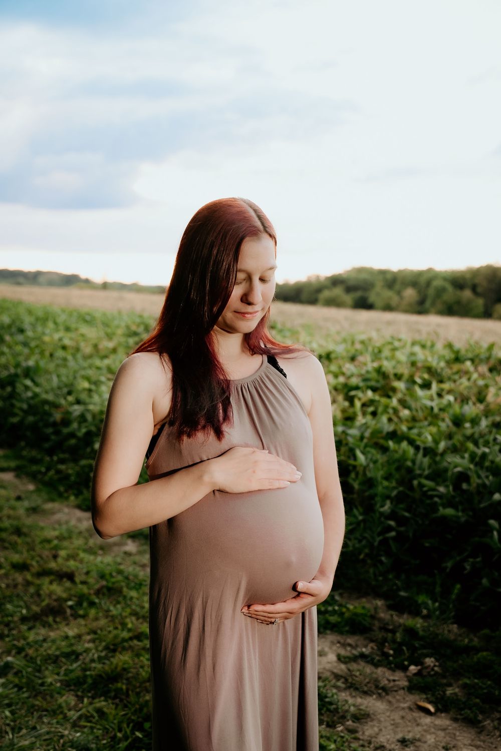 Expectant mother in taupe dress poses in sunlit field.