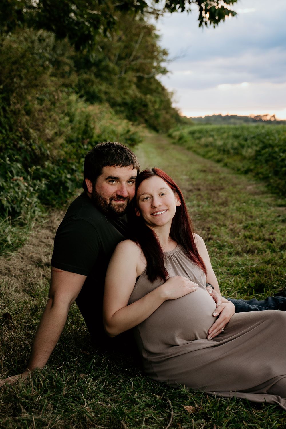 Intimate maternity photoshoot on grassy hillside showing couple in casual attire embracing outdoors.