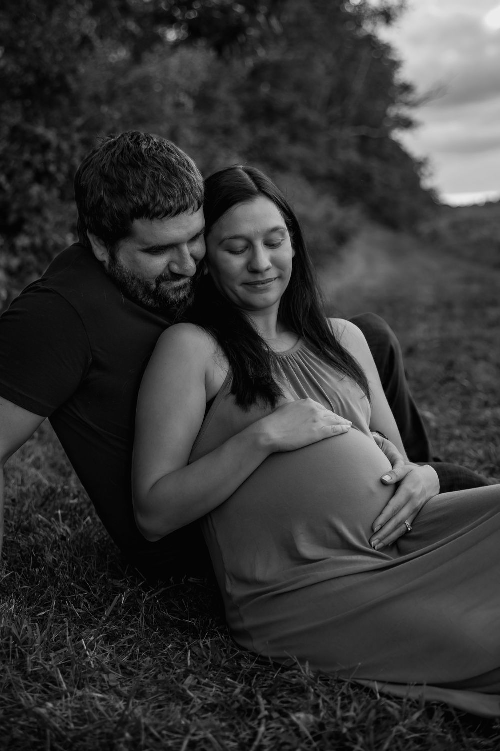 Black and white maternity photos showing tender moments between couple on grassy hill.