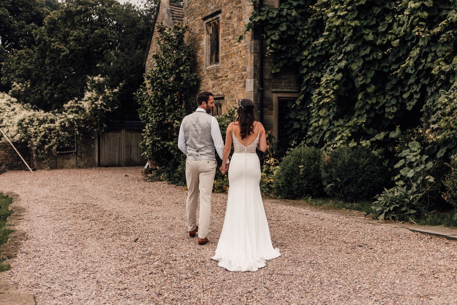 A couple walks down a gravel path next to a historic stone building surrounded by lush greenery.