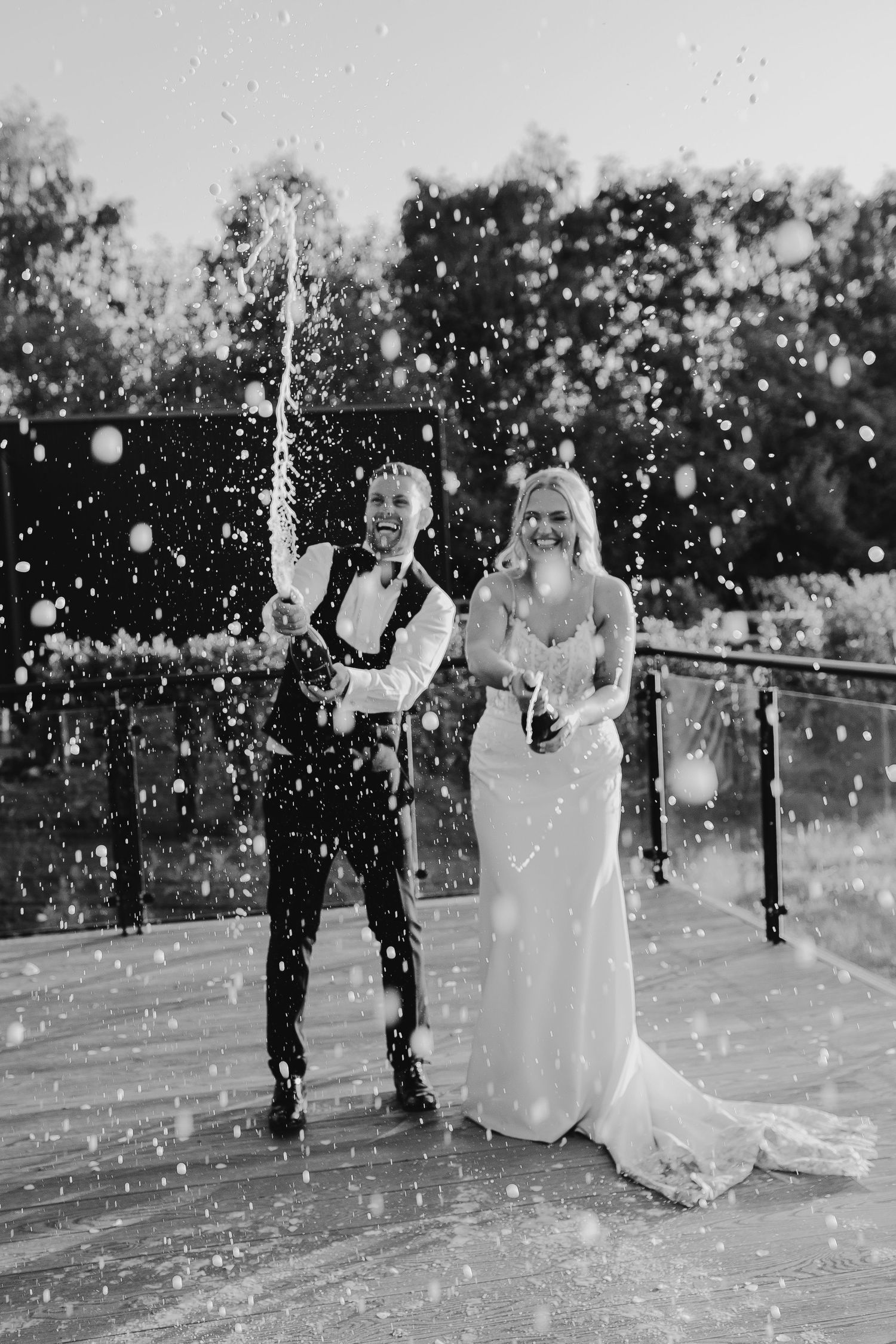 Black and white photo of a joyful bride and groom popping champagne outdoors, with bubbles spraying everywhere.