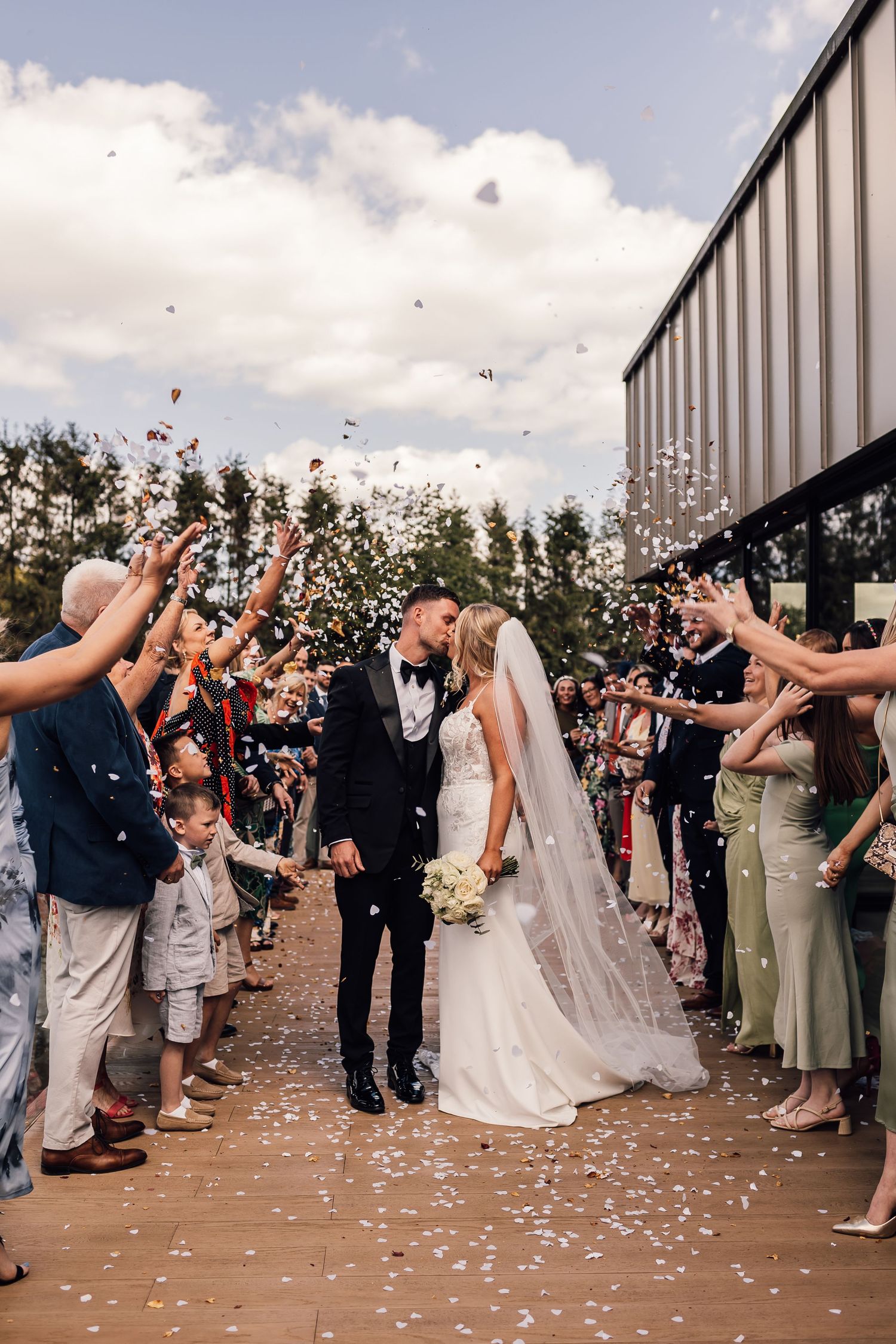 Bride and groom kissing during confetti exit at outdoor wedding ceremony with guests celebrating.