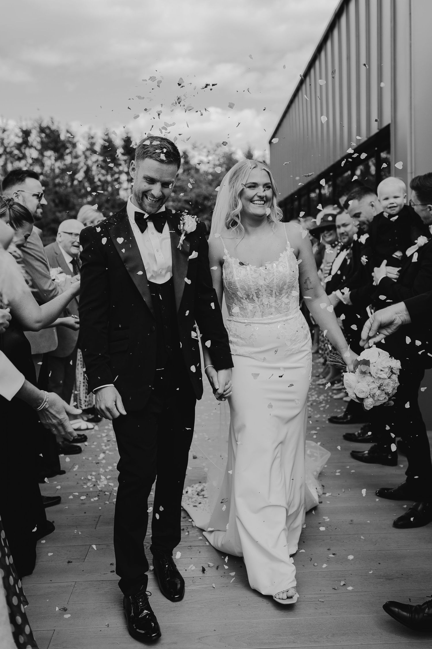 Black and white photo of a smiling bride and groom walking through a confetti shower at their wedding ceremony.