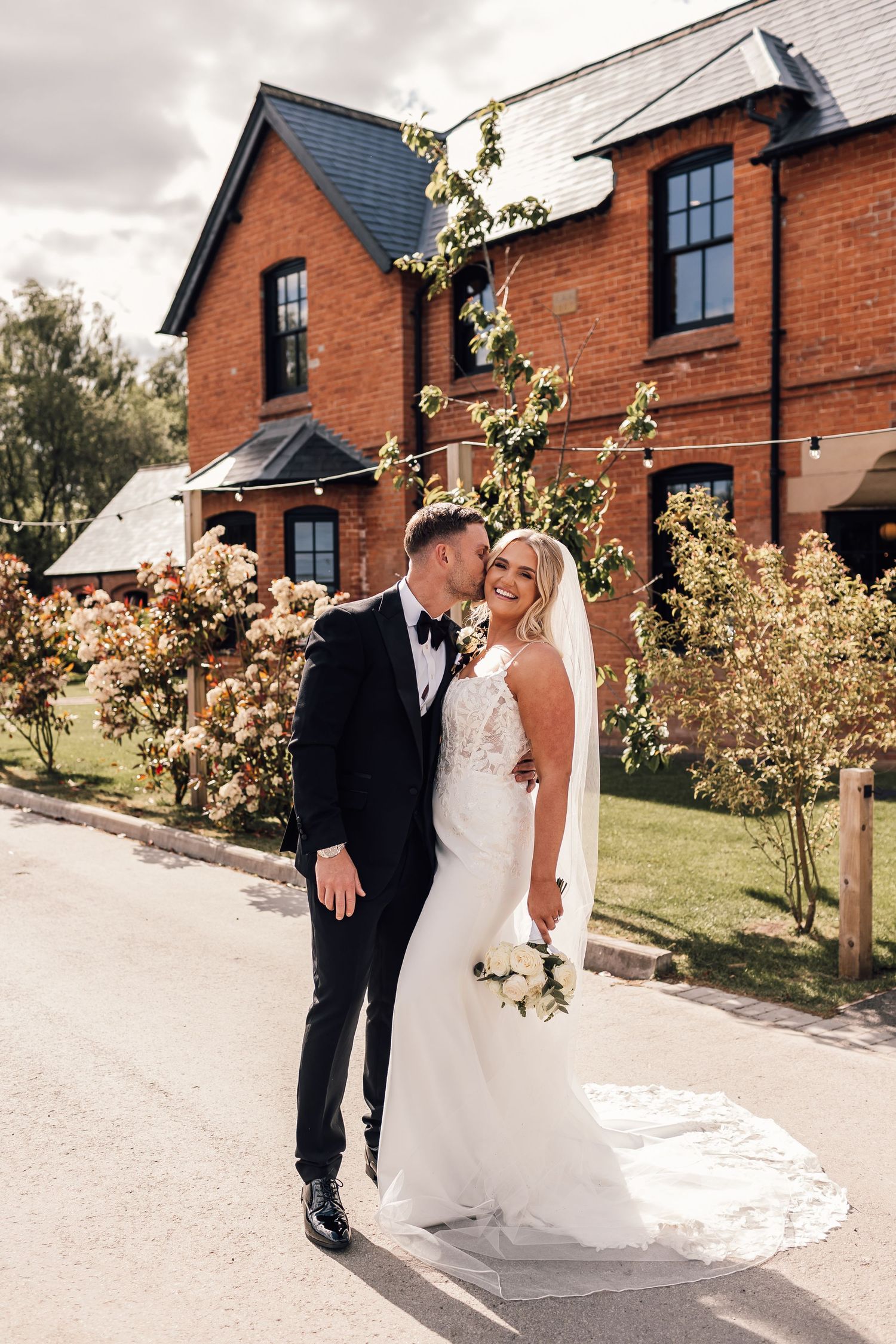 Bride and groom posing outside a red brick wedding venue surrounded by blooming flowers on their wedding day.