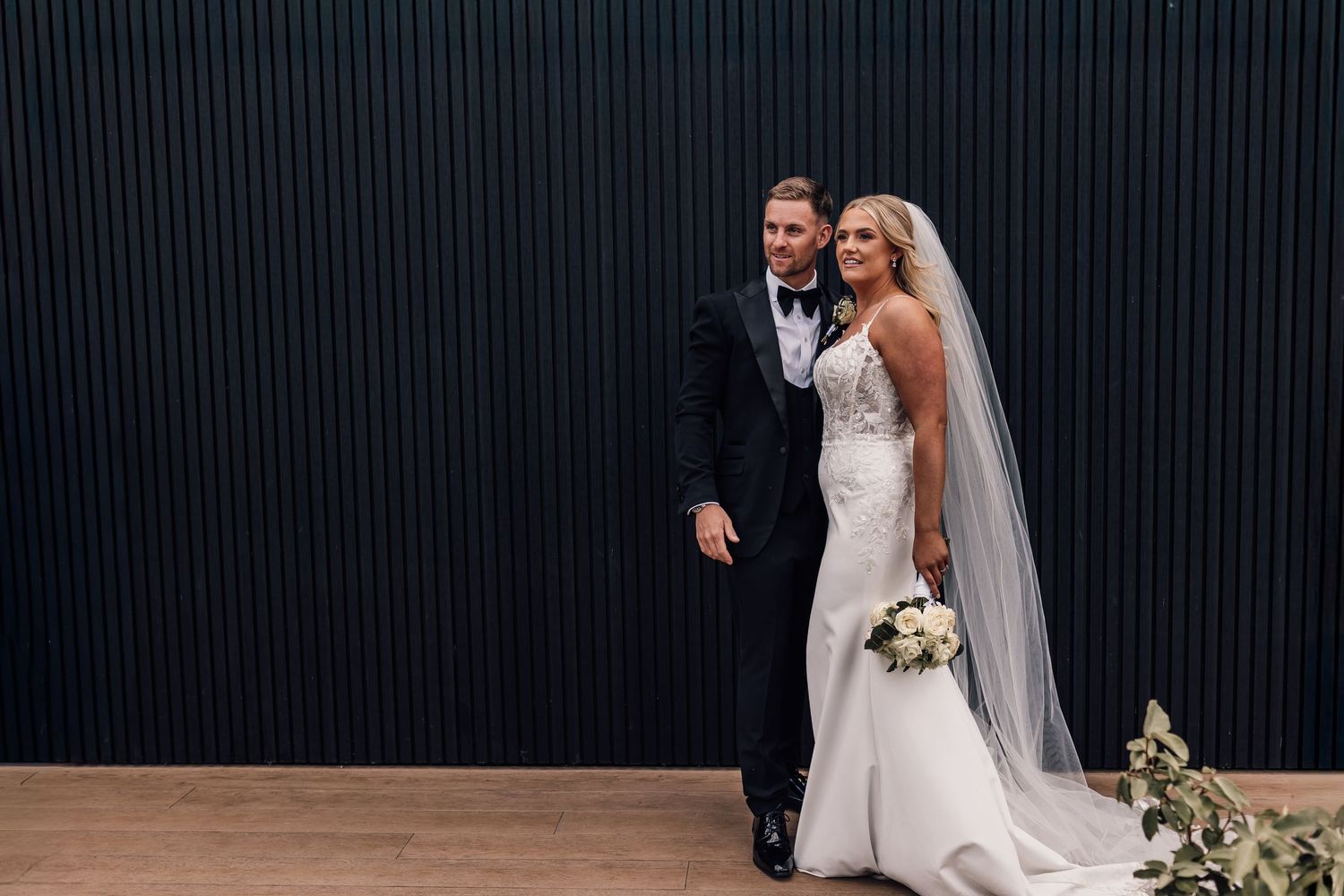 Bride in white mermaid gown with veil and groom in black tuxedo posing together against a dark wall.
