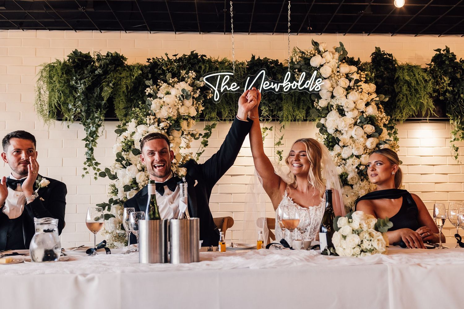Bride and groom raise hands in celebration at elegant wedding reception with floral backdrop and The Newbolds neon sign.