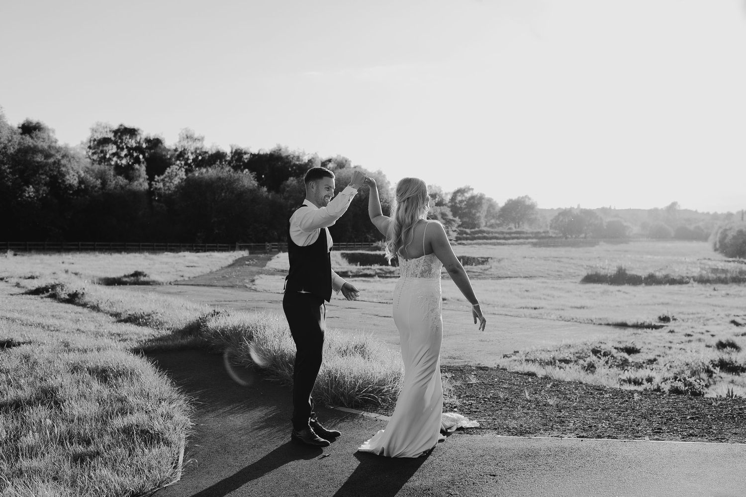 Black and white photo of a bride and groom dancing outdoors on a sunny day with scenic fields in the background.