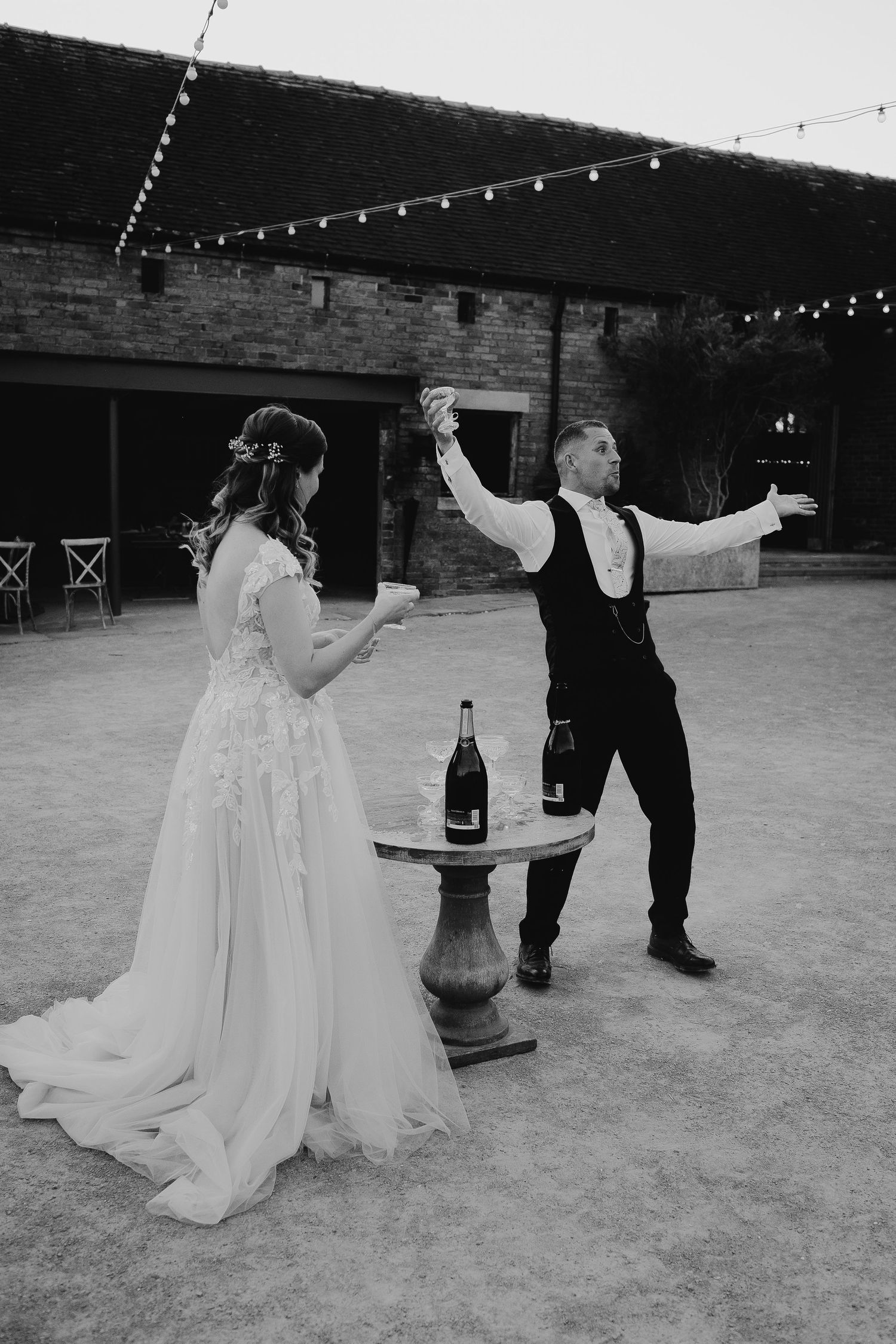 Black and white photo of bride and groom dancing outdoors at wedding reception with string lights and champagne table.